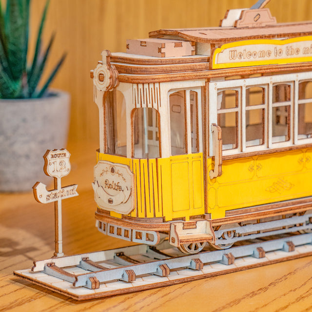 Wooden ROBOTIME Rolife kits Tramcar on a wooden surface with a blurred plant in the background